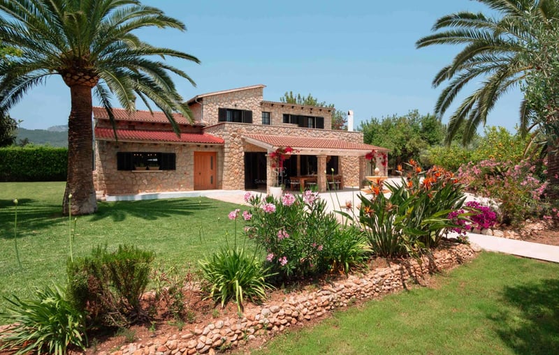 Villa en pierre naturelle de deux étages avec toits en terre cuite, palmiers et fleurs éclatantes par temps clair.
