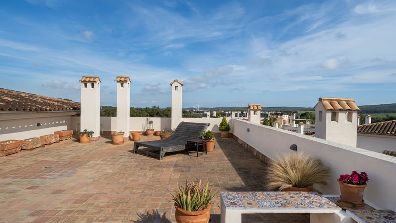 Grande terrasse de toit avec carrelage en terre cuite, cheminées blanches et chaise longue en bois sous un ciel bleu.