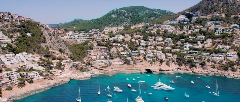 Vue en plongée de maisons blanches sur une colline verdoyante surplombant une baie avec des yachts.