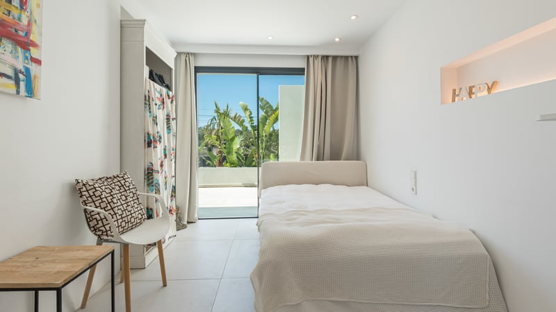 Chambre moderne blanche avec lit simple, grandes baies vitrées donnant sur une terrasse et végétation tropicale.