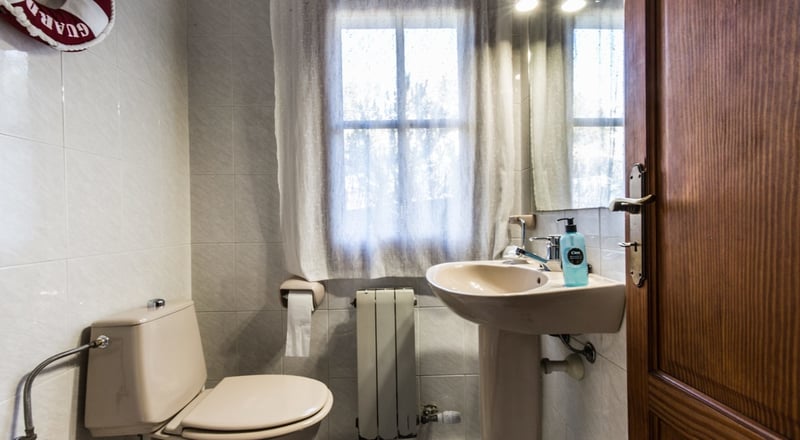 Petite salle de bain avec toilettes et lavabo colonne beige, fenêtre avec rideau et murs carrelés clairs.