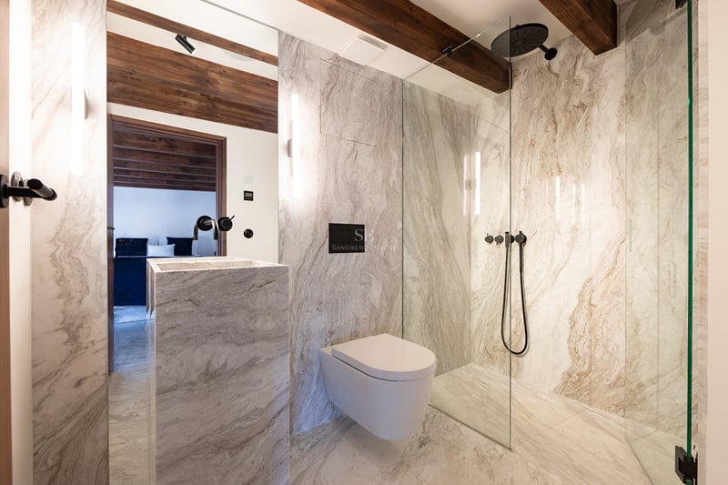 Baño moderno con paredes de mármol, ducha de cristal, grifería negra mate y vigas de madera vista en el techo.