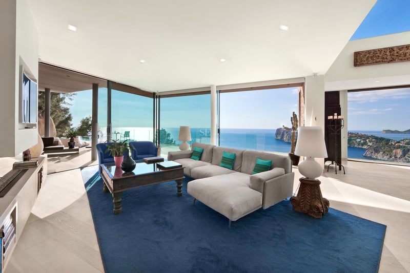 Salon moderne avec canapé gris, tapis bleu et grandes baies vitrées offrant une vue imprenable sur l'océan.