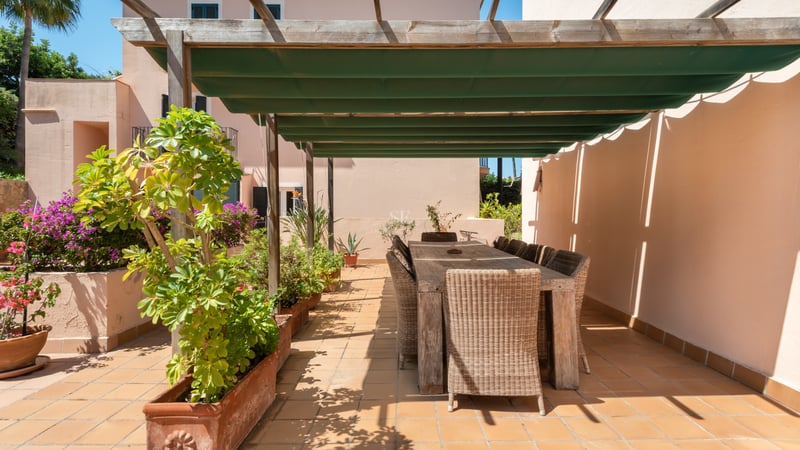 Espace repas extérieur sur une terrasse en terre cuite avec pergola en bois, store vert et chaises en osier.