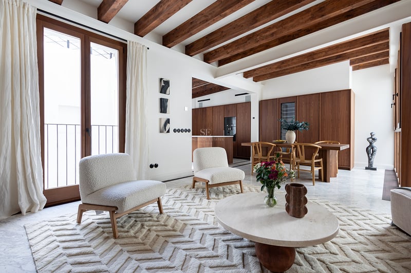 Sala de estar moderna con vigas de madera oscura, sillones bouclé blancos, suelo de mármol y comedor al fondo.