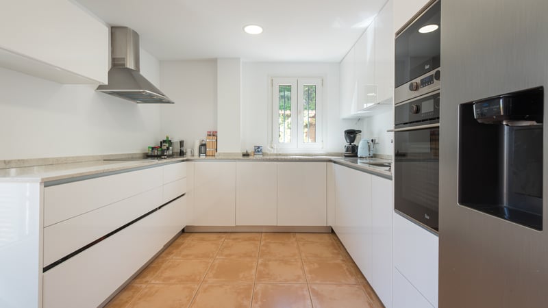 Cuisine moderne en U avec placards blancs brillants, sol en terre cuite et appareils en acier inoxydable.