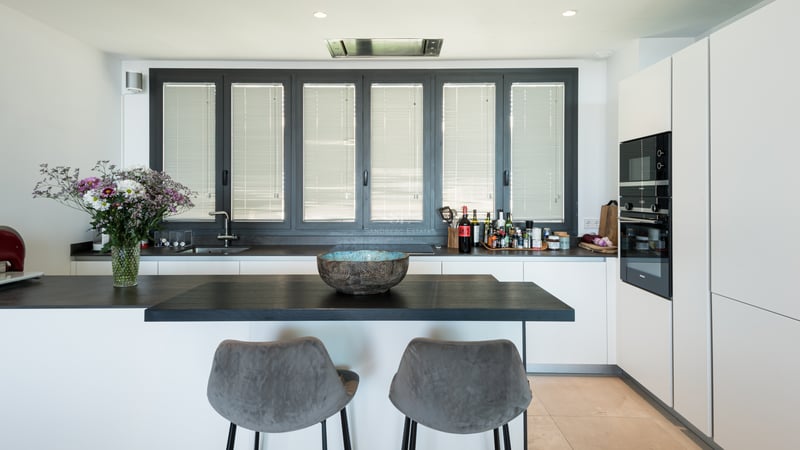 Cuisine blanche moderne avec plans de travail sombres, îlot avec deux tabourets gris et grandes fenêtres.