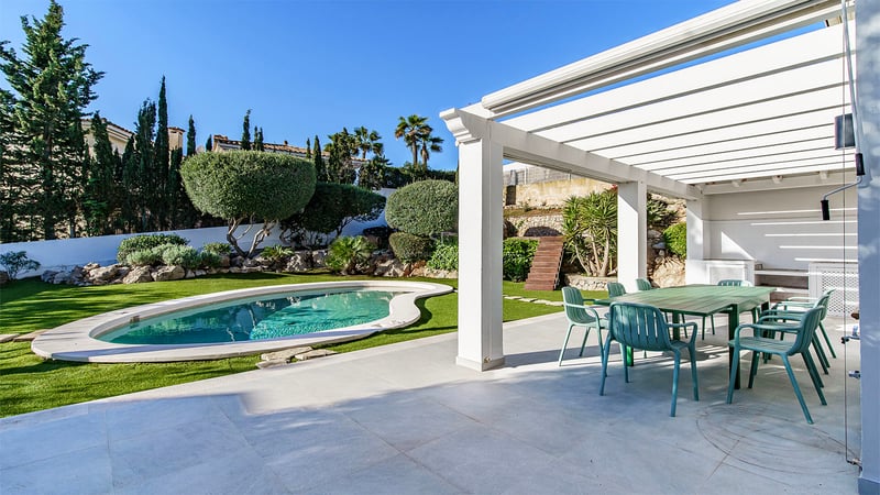 Pergola blanche abritant un coin repas extérieur avec chaises vertes à côté d'une piscine et d'un jardin paysager.
