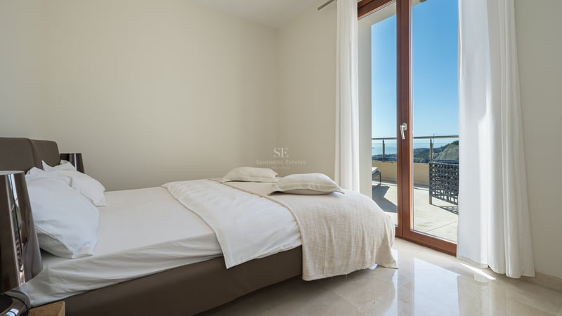 Chambre moderne avec draps blancs, sol en marbre poli et grandes baies vitrées ouvrant sur une terrasse ensoleillée.