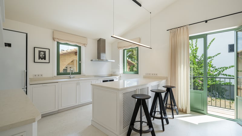 Cuisine blanche moderne avec îlot central, tabourets noirs et fenêtres à cadres verts sous une lumière naturelle.