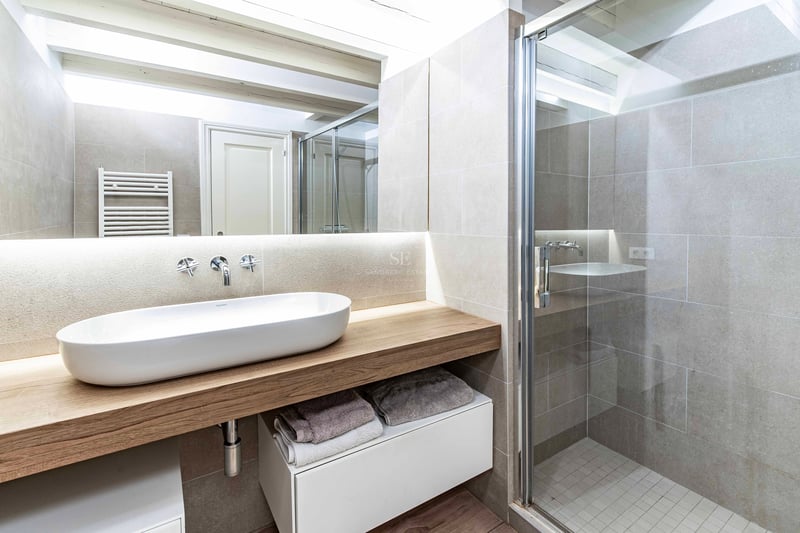 Salle de bains avec vasque ovale sur plan de travail en bois, douche vitrée et poutres apparentes blanches.