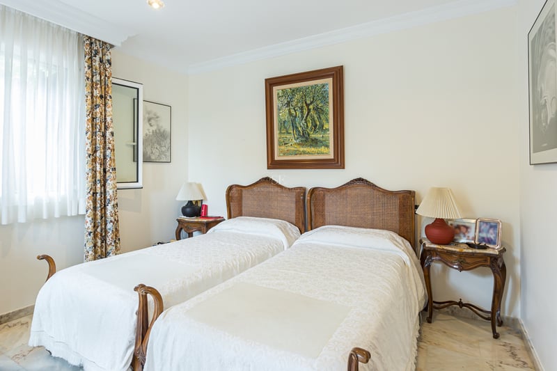 Chambre avec deux lits simples, têtes de lit en rotin, sol en marbre et tableaux au mur.