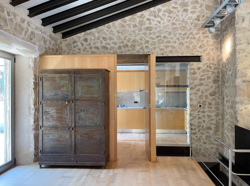 Intérieur avec murs en pierre naturelle, parquet en bois clair, poutres foncées et vue sur une cuisine moderne.