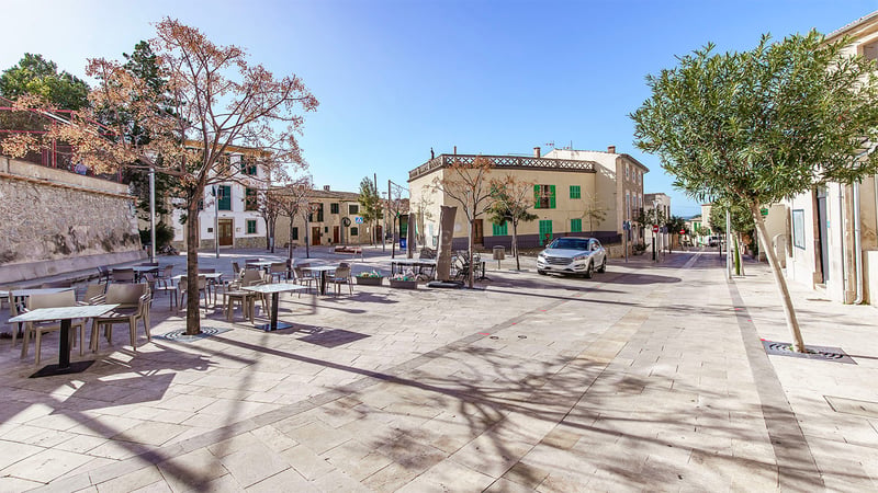 Place ensoleillée avec terrasses de café, architecture traditionnelle en pierre et arbres sous un ciel bleu pur.