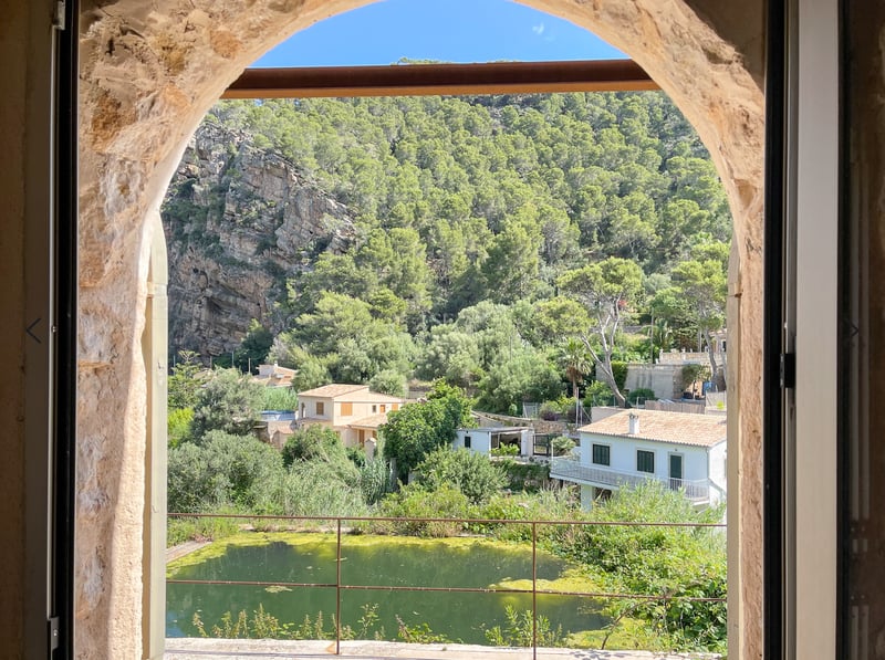 Vue à travers une arche en pierre sur une vallée verdoyante, des falaises rocheuses et des maisons traditionnelles.