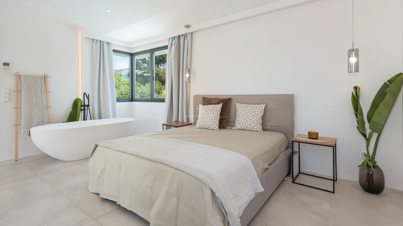 Chambre minimaliste avec baignoire blanche, literie neutre et fenêtres d'angle donnant sur la verdure.