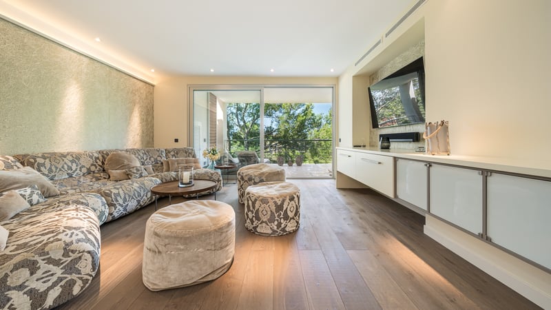 Vaste salon avec canapé à motifs, parquet en chêne et grandes baies vitrées ouvrant sur une terrasse boisée.
