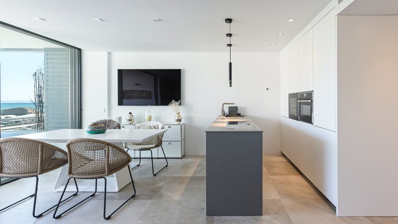 Cocina blanca moderna con isla gris, mesa de comedor y grandes ventanales con vistas al Mediterráneo.