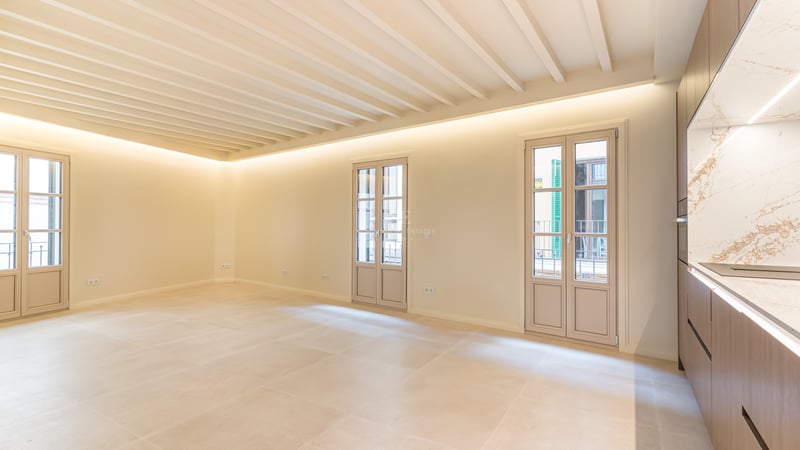 An empty, bright living room with cream tile floors, a beamed ceiling, three French doors, and a modern kitchen.