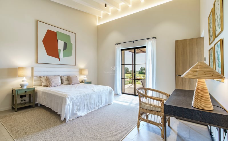 Dormitorio luminoso con techos altos, cama blanca, silla de mimbre y grandes puertas de cristal al jardín.