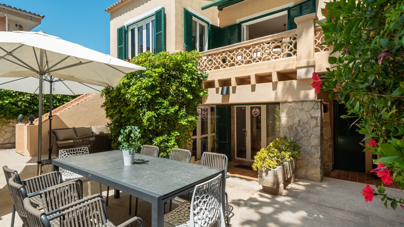 Terrasse en pierre avec salle à manger extérieure grise, parasol blanc et façade de villa aux volets verts.