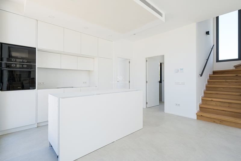 Cuisine moderne blanche avec îlot central, appareils intégrés et escalier en bois menant à l'étage.