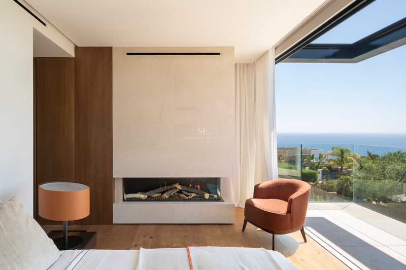 Chambre de maître moderne avec cheminée en pierre, fauteuil orange et baies vitrées ouvrant sur une terrasse vue mer.