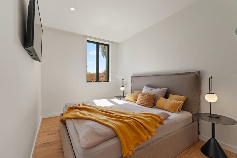Chambre moderne avec lit gris, jeté de lit moutarde, parquet en bois et fenêtre lumineuse.