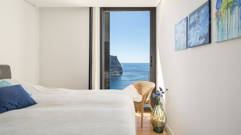 Chambre lumineuse avec literie blanche, oreiller bleu et baies vitrées ouvrant sur un paysage côtier méditerranéen.