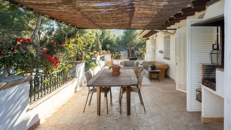 Terrasse ombragée avec table à manger en bois, meubles en osier et barbecue intégré entourée de pins.