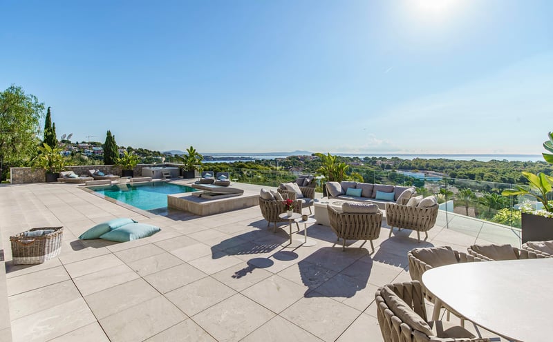 Terrasse en pierre avec mobilier design, piscine et vue panoramique sur la mer et la verdure par grand soleil.