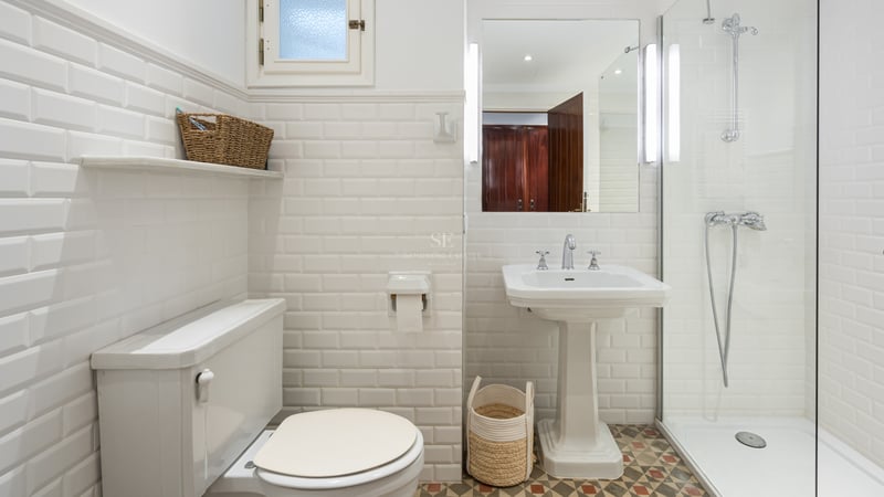 Salle de bain lumineuse avec carrelage métro blanc, lavabo sur colonne, douche vitrée et sol à motifs.