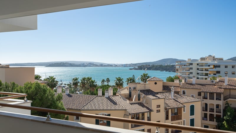 Vista desde un balcón hacia tejados mediterráneos, palmeras y una amplia bahía turquesa bajo un cielo despejado.