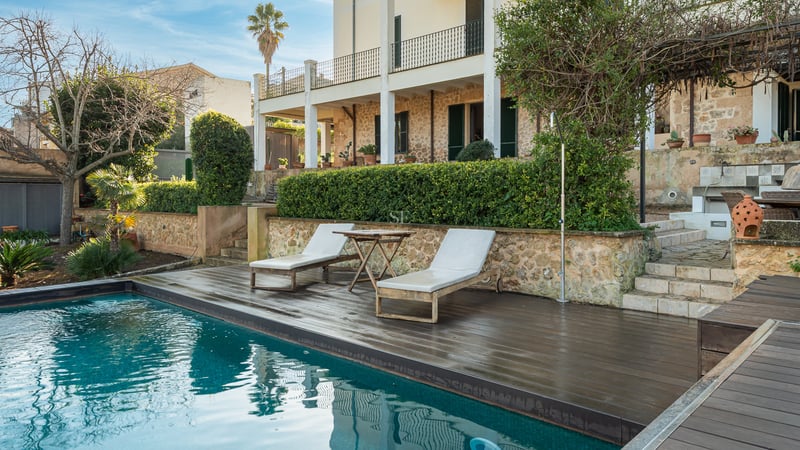 Piscine turquoise avec terrasse en bois sombre, chaises longues et façade en pierre rustique.