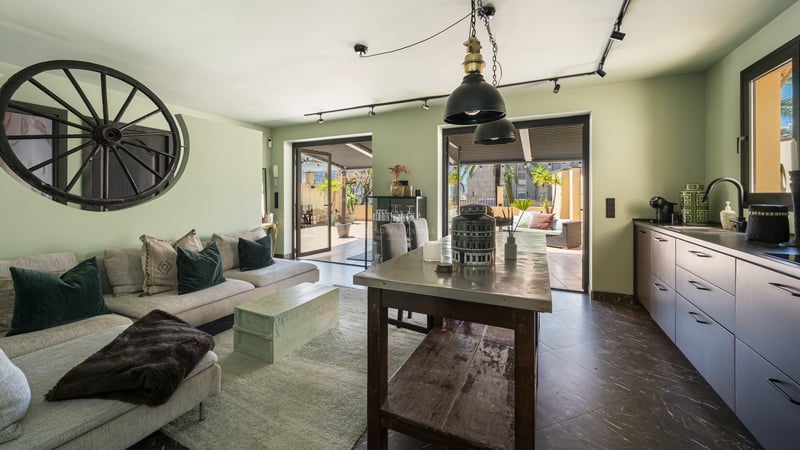 Intérieur d'un salon vert sauge et cuisine design s'ouvrant sur une terrasse privée ensoleillée.