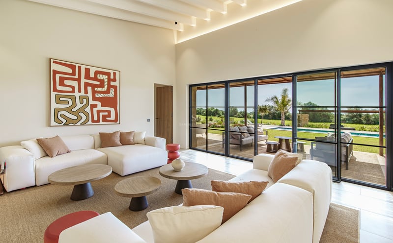 Salón luminoso con sofás blancos, mesas de madera y grandes ventanales con vistas al jardín y la piscina.