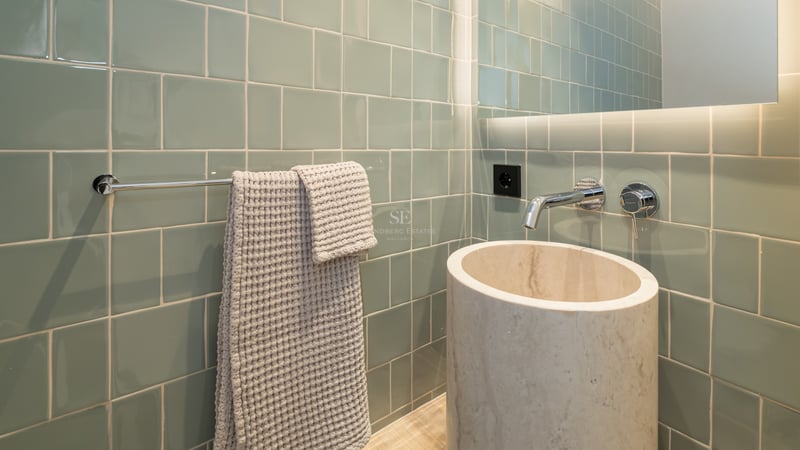 Vasque cylindrique en marbre crème devant des carreaux vert sauge avec robinetterie chromée encastrée.
