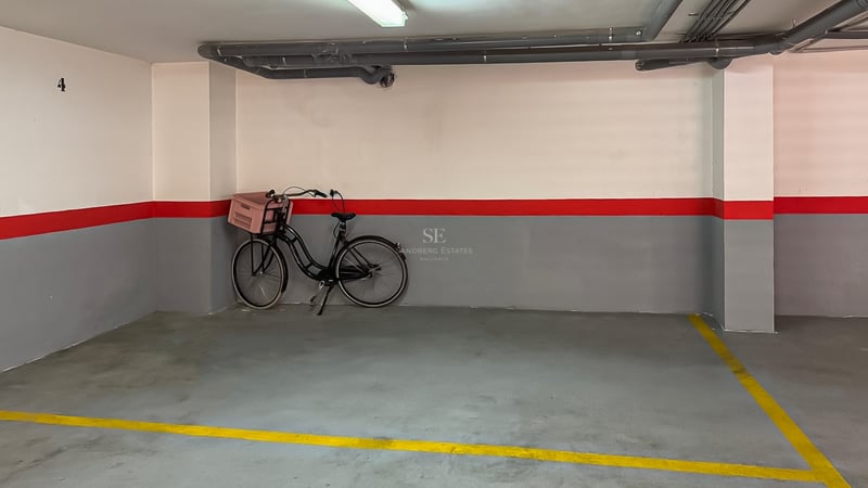 Place de parking numérotée avec murs gris et blancs, bande rouge et sol en béton avec lignes jaunes.