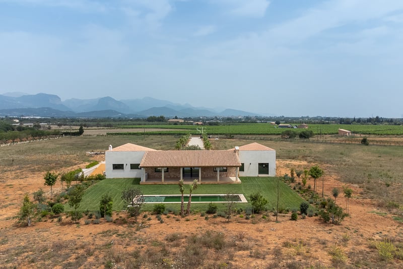 Vue aérienne d'une villa blanche avec piscine, pelouse verte et montagnes au loin sous un ciel voilé.