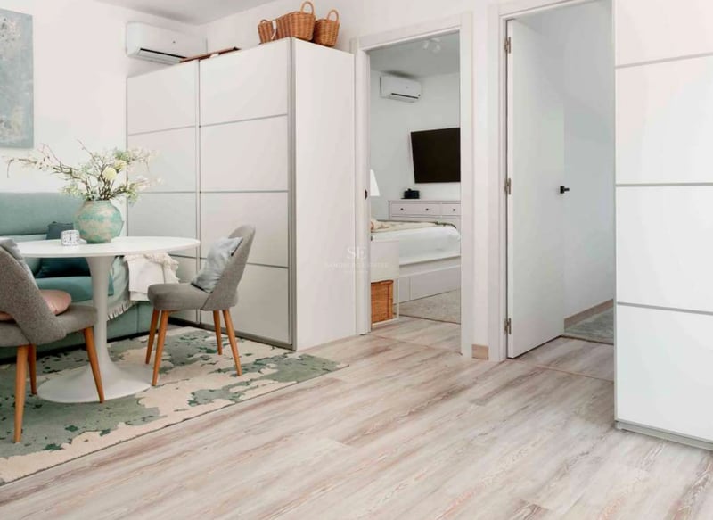Espace de vie lumineux avec table ronde blanche, chaises grises, parquet clair et vue sur la chambre attenante.